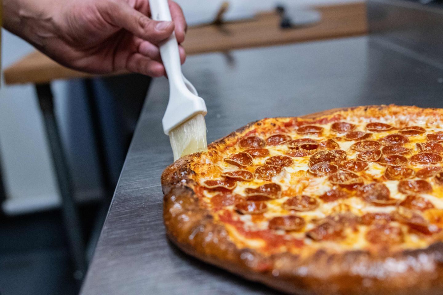 Hand brushing pizza crust with oil on a metal countertop.