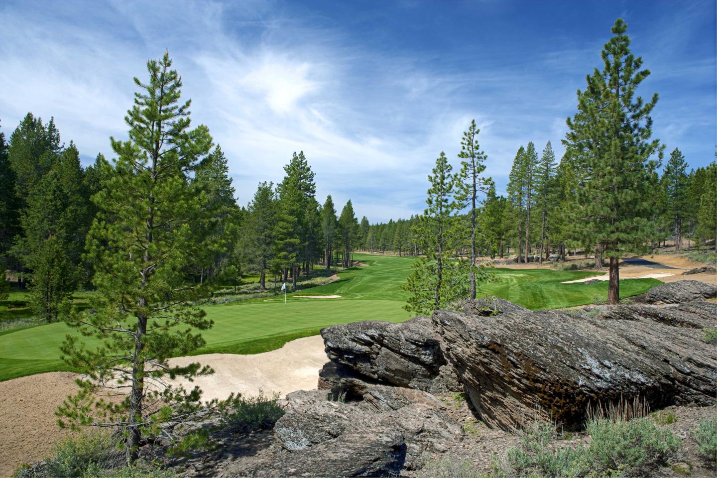 Golf course surrounded by pine trees and rocky terrain under a blue sky.