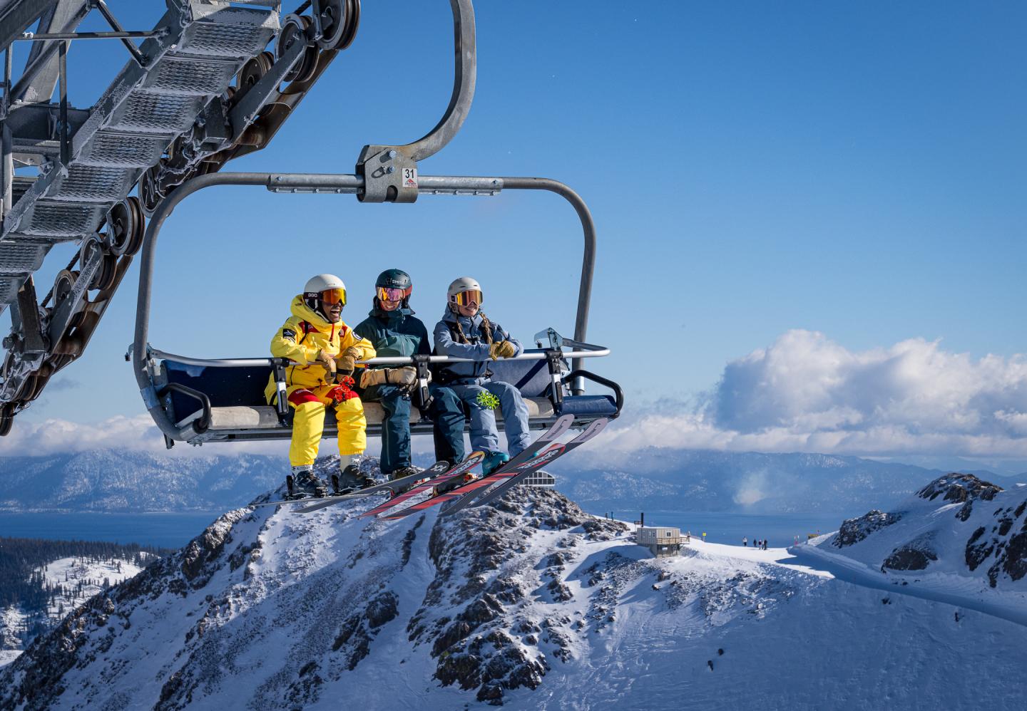 Three skiers on a chairlift over snowy mountains, clear blue sky.