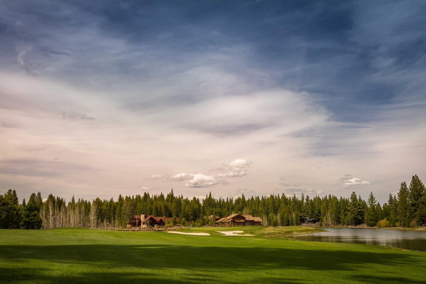 Green field with trees, distant cabin, under a partly cloudy sky.