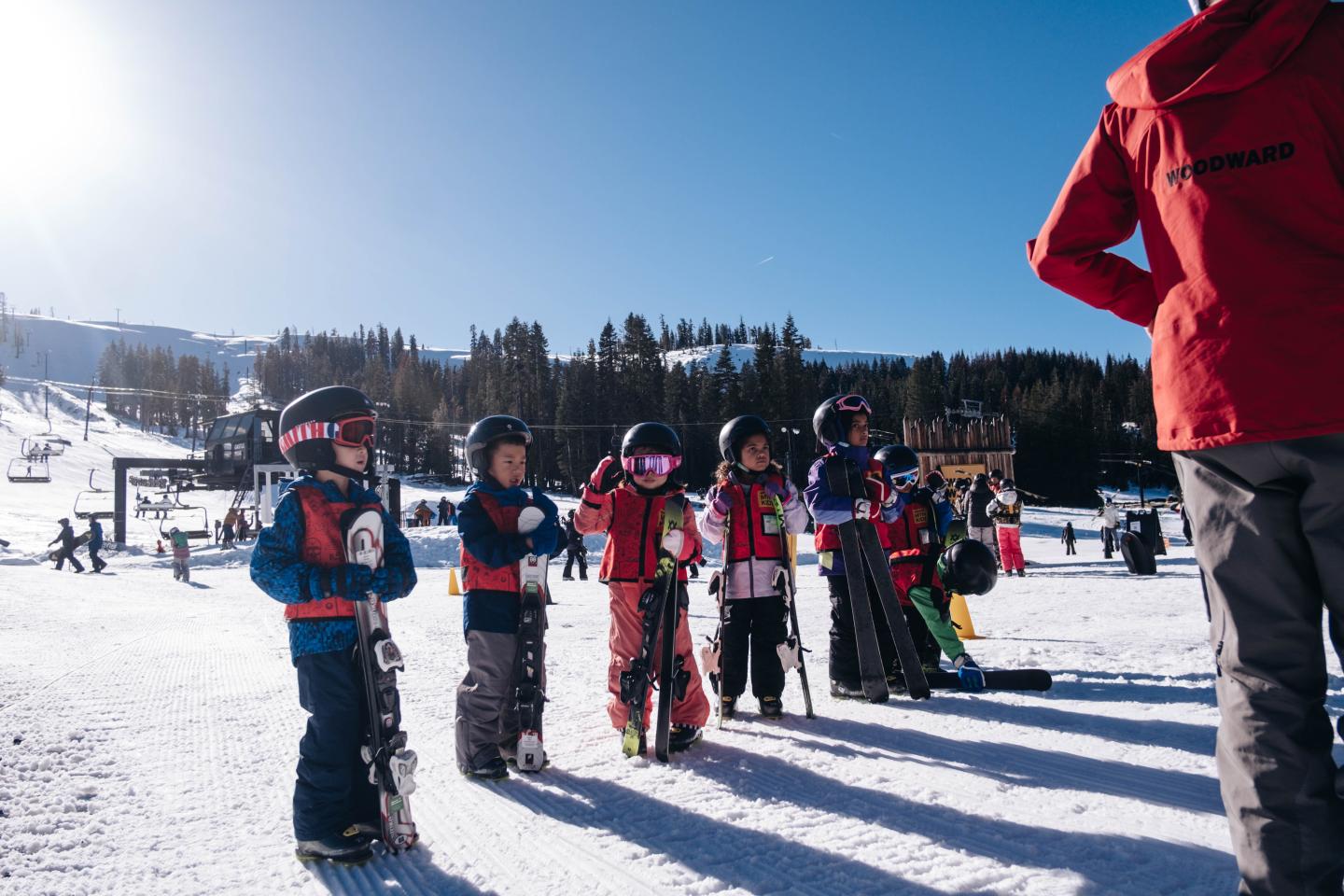Children in ski gear stand in a snowy area under a clear blue sky.