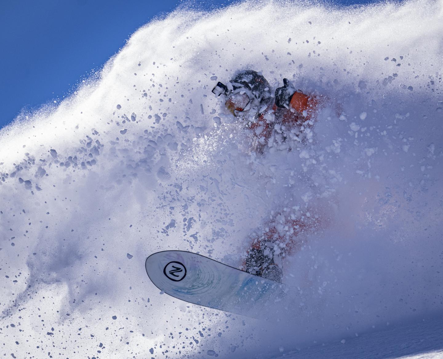 Snowboarder riding through powder under a clear blue sky.