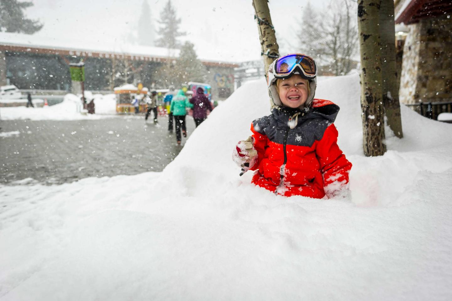 Child in red snowsuit playing in snow, wearing goggles, with snowy backdrop.