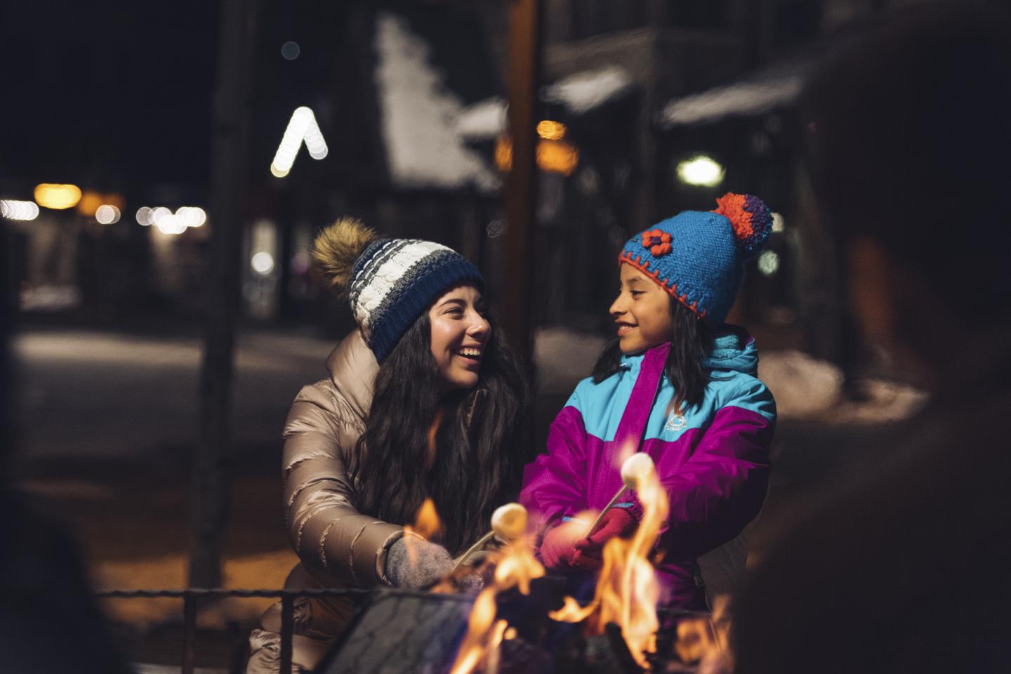 Two people in winter clothes smiling by a campfire at night.