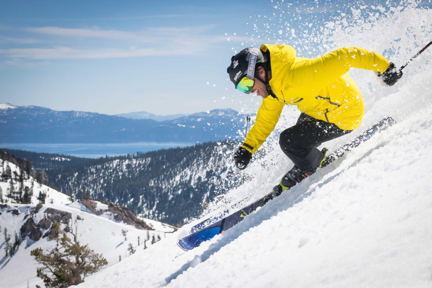 Skier in yellow jacket descends snowy slope, with mountains and lake in background.