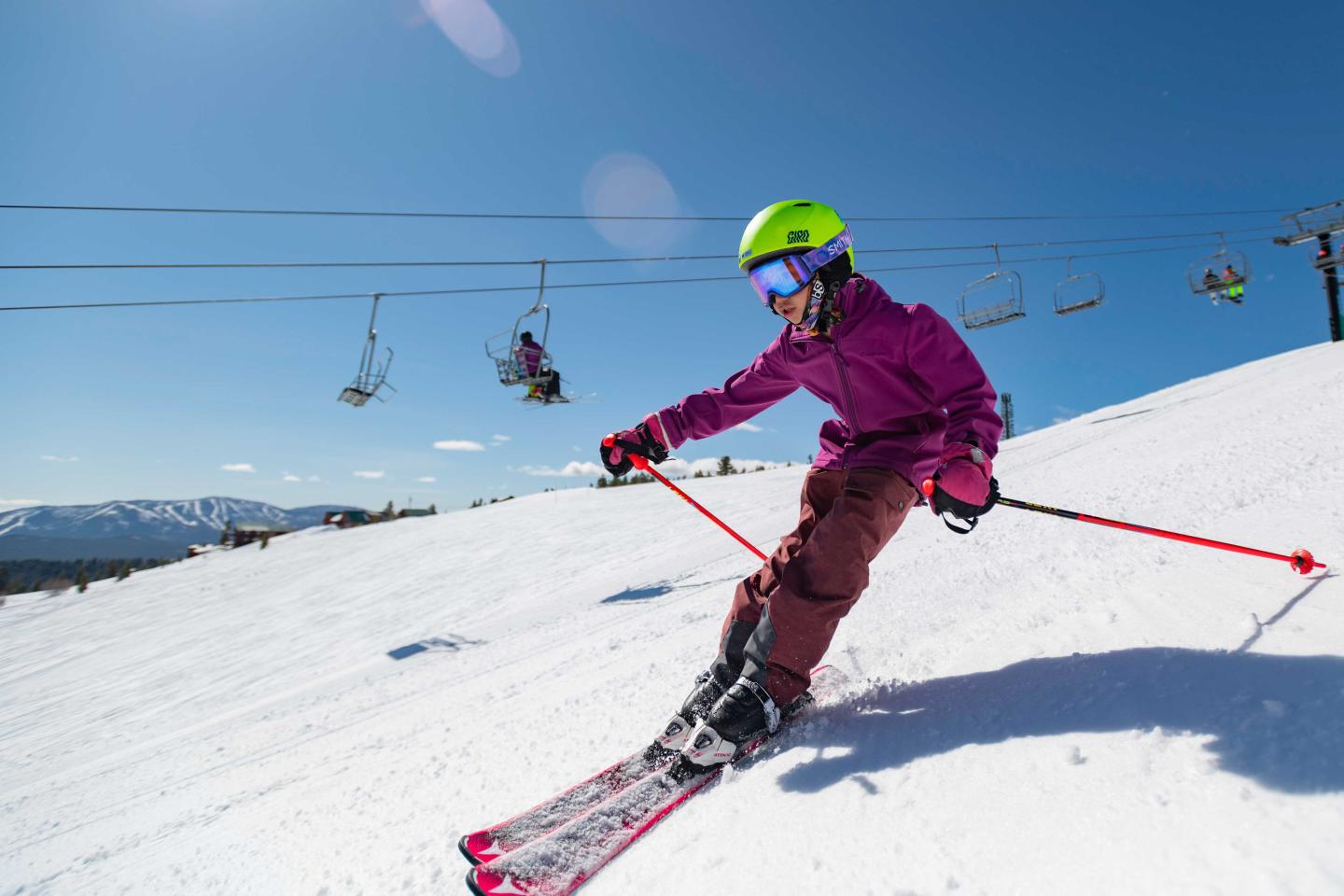 Skier in purple jacket and green helmet descends a snowy slope under blue sky.