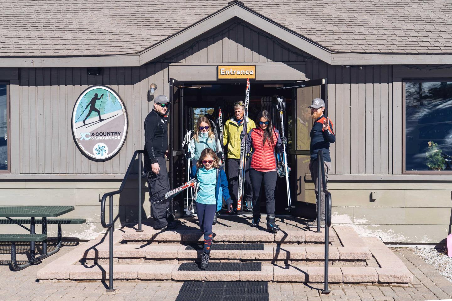 Skiers exit a lodge on a sunny day, carrying skiing gear.