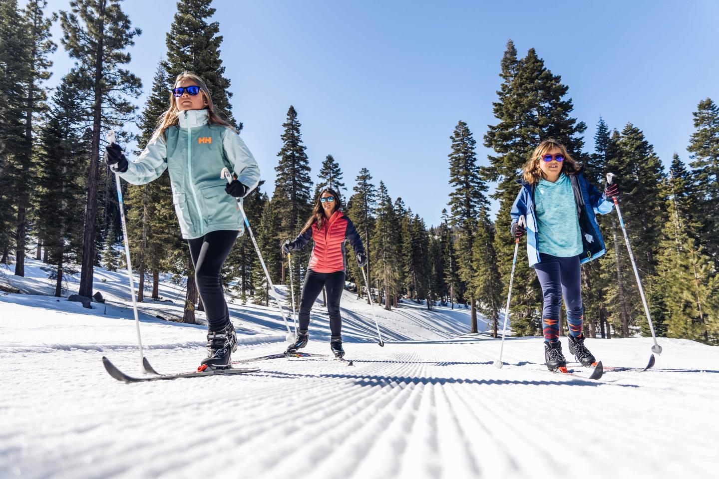 Three people cross-country skiing in a snowy forest under a clear blue sky.