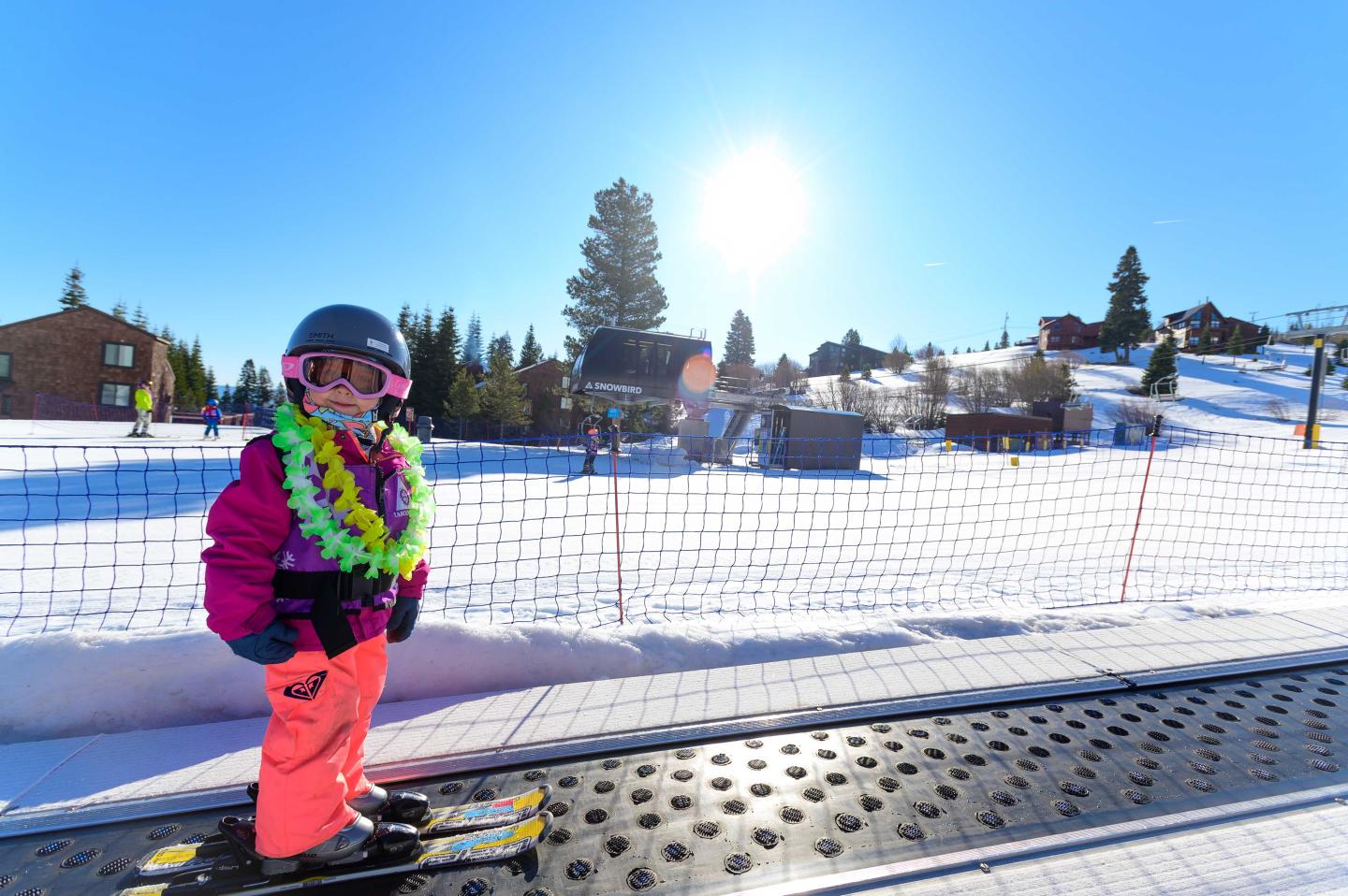 Child in ski gear with a floral lei, on a snowy mountain slope under bright sun.