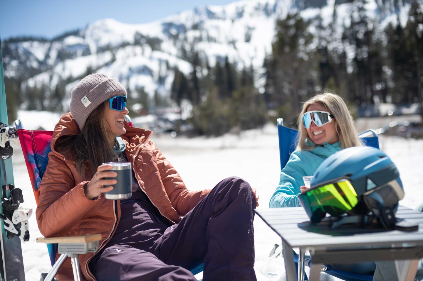 Two people in winter gear relax outdoors with snowy mountains in the background.