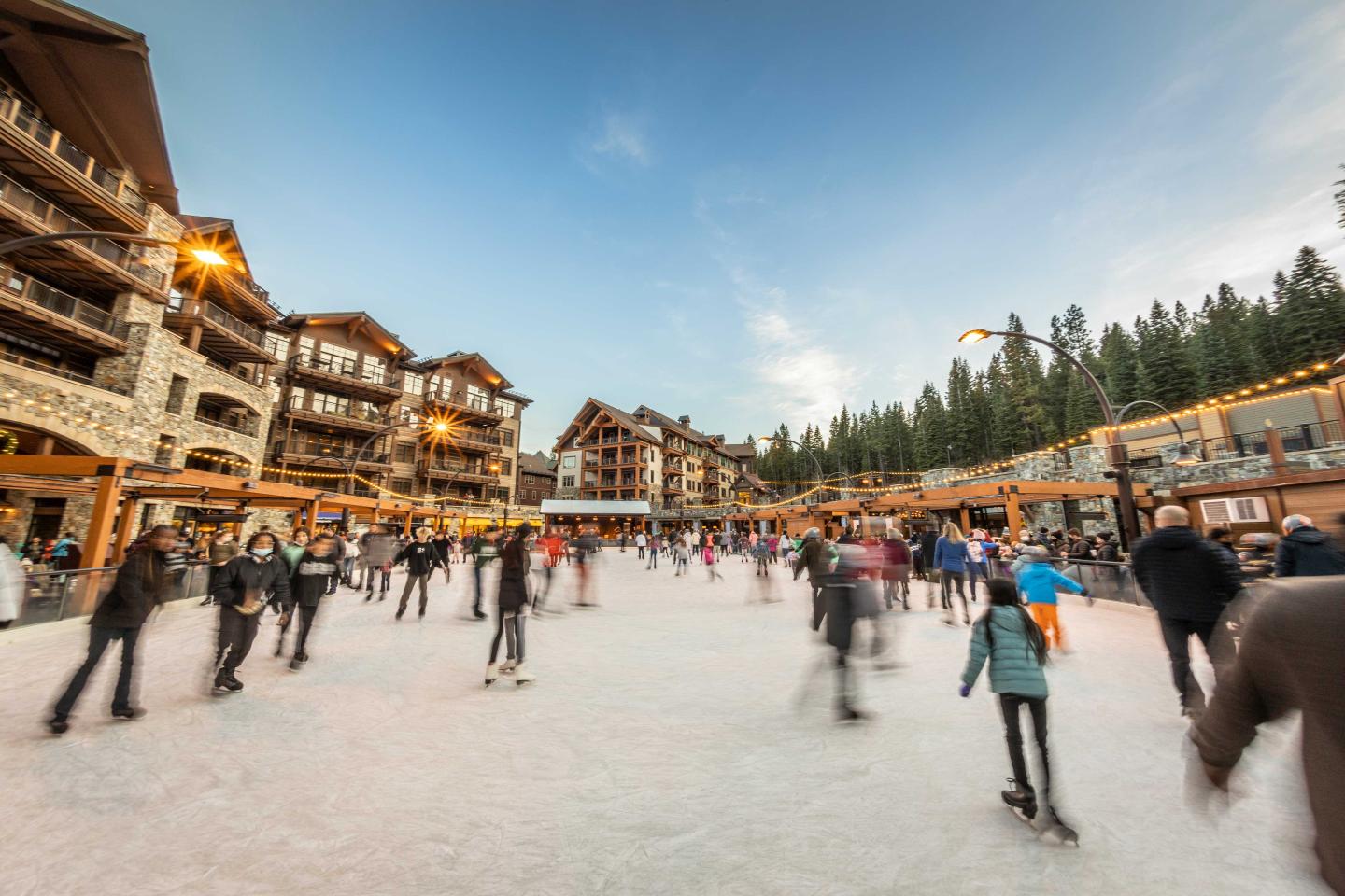 People ice skating in a vibrant outdoor rink surrounded by lodges and trees.