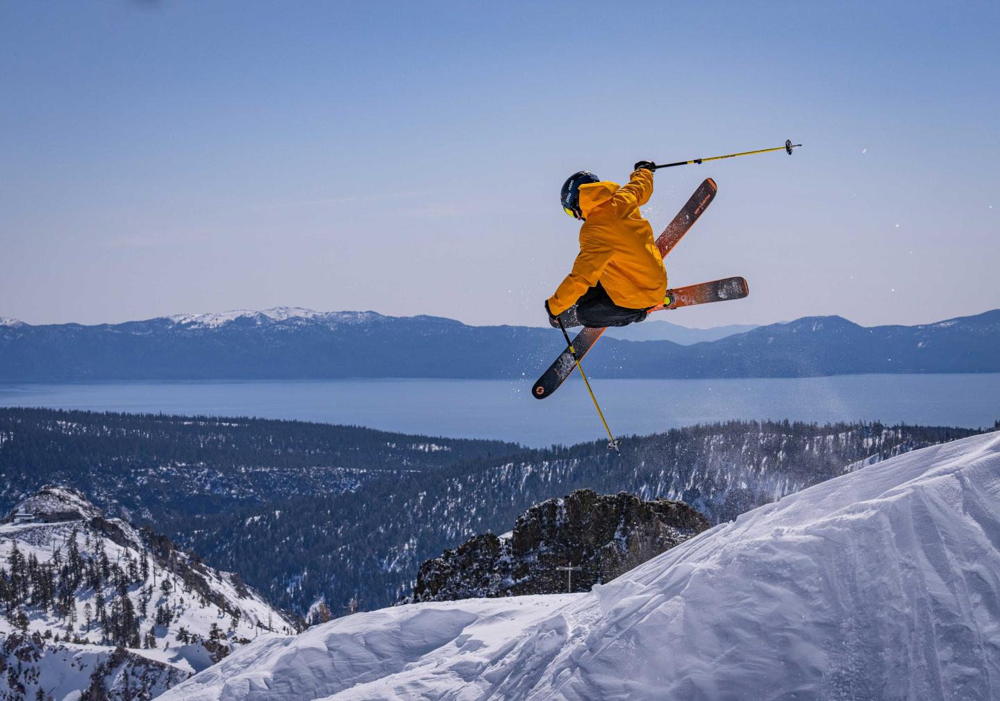 Skier in orange jacket jumps off snowy mountain with lake view in background.