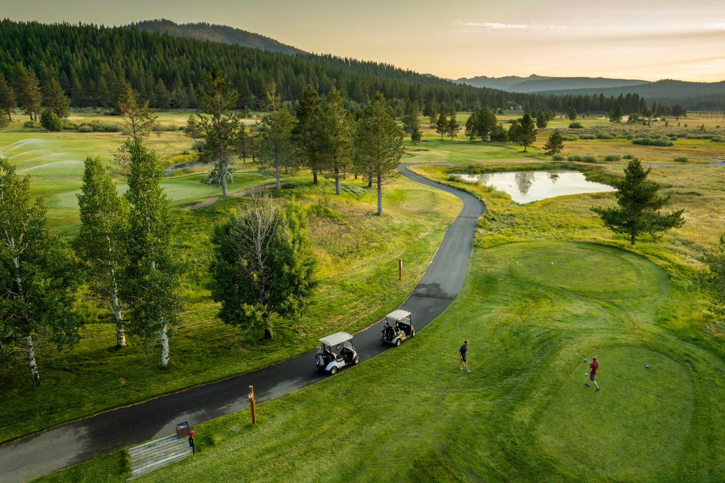 Golf course at sunset with two carts, lush greenery, and distant mountains.