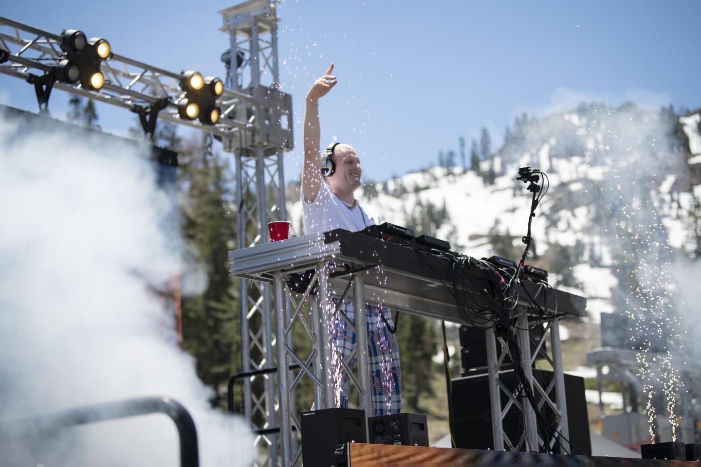 DJ performing on an outdoor stage with snow-covered mountains in the background. Spring Tracks Concert Series at Palisades Tahoe.
