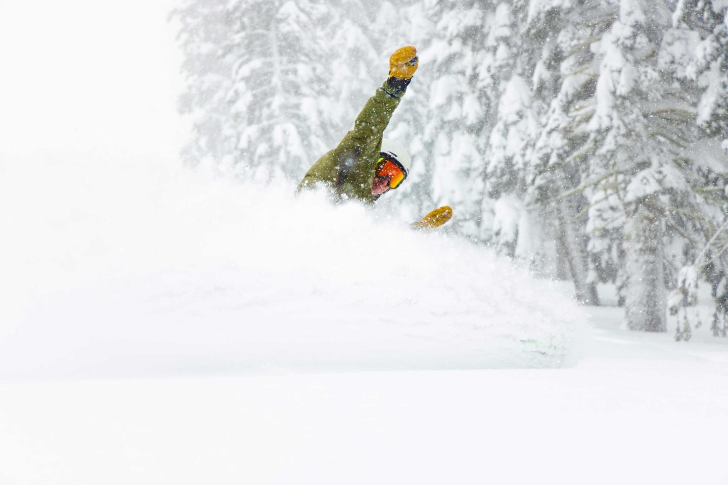 Snowboarder in green gear riding through deep powder snow, surrounded by snowy trees.