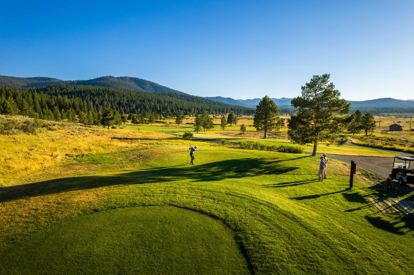 Golfers on a lush, sunny course with mountains in the background.