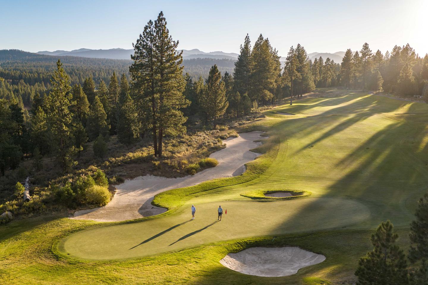 Golf course with two players, surrounded by tall trees at sunset.