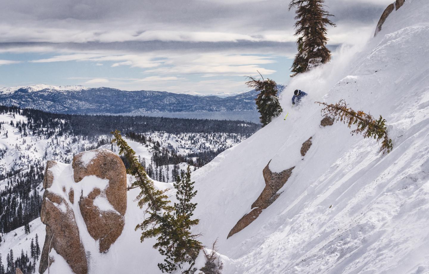 Snowboarder descends snowy mountain slope with pine trees, mountains in background.