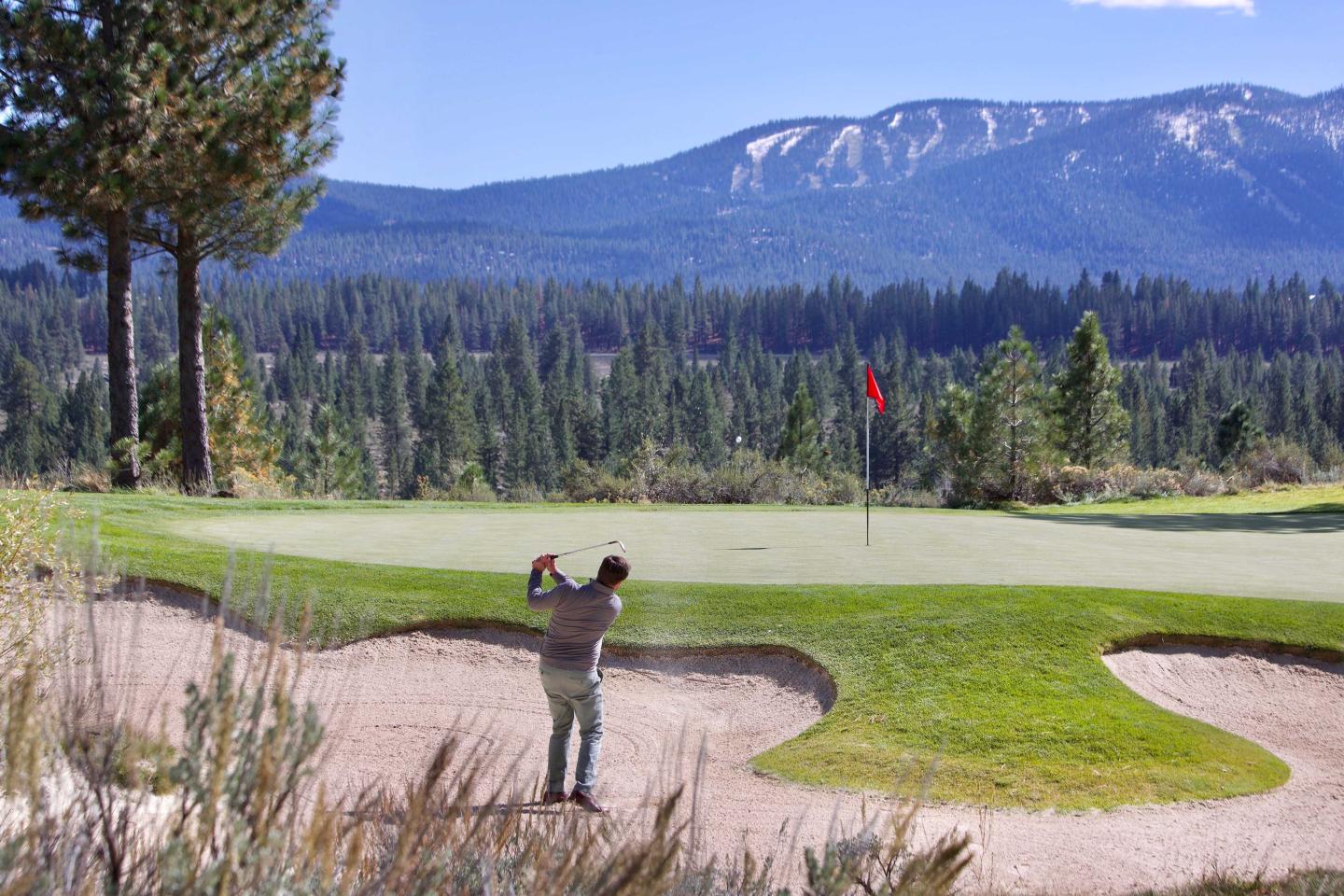 Golfer swings on a sunny course with mountains in the background.