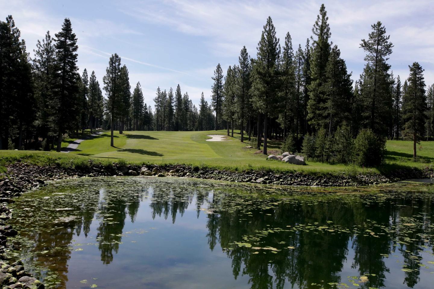 Golf course with green fairway, surrounded by tall pines, reflected in a pond under a blue sky.