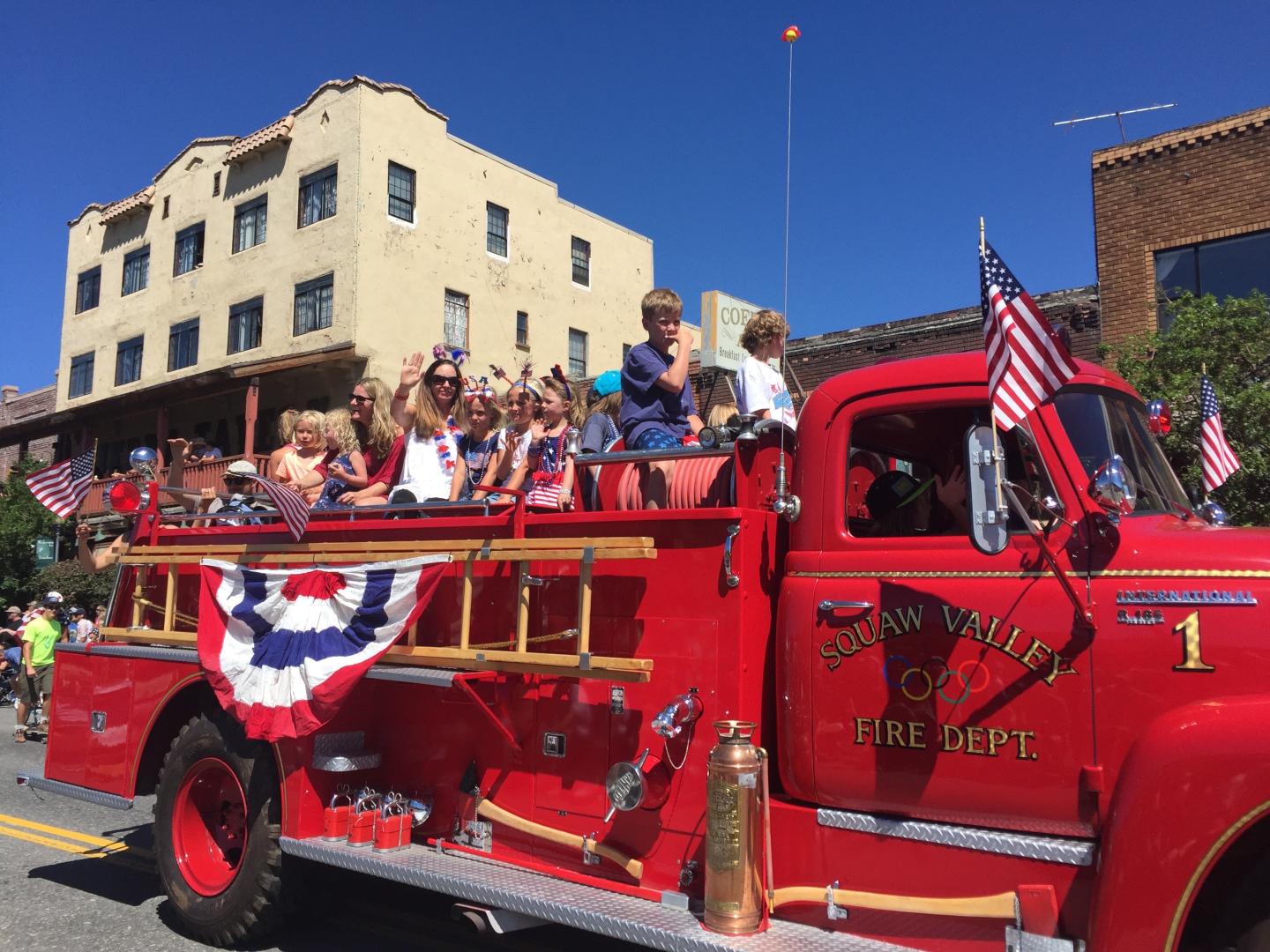 Red vintage firetruck in a parade, decorated with flags and bunting, children waving.