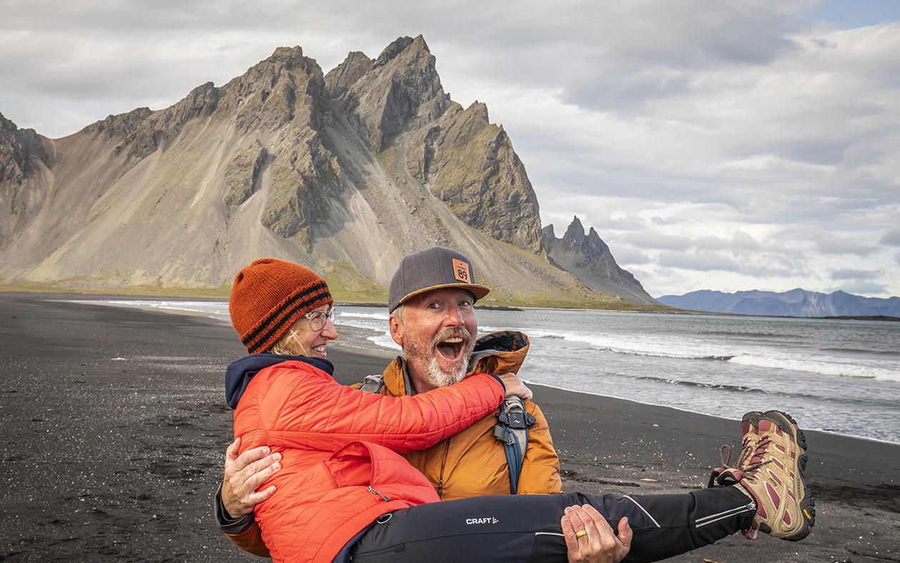 Elderly man joyfully carrying a woman on a black sand beach with mountains behind.