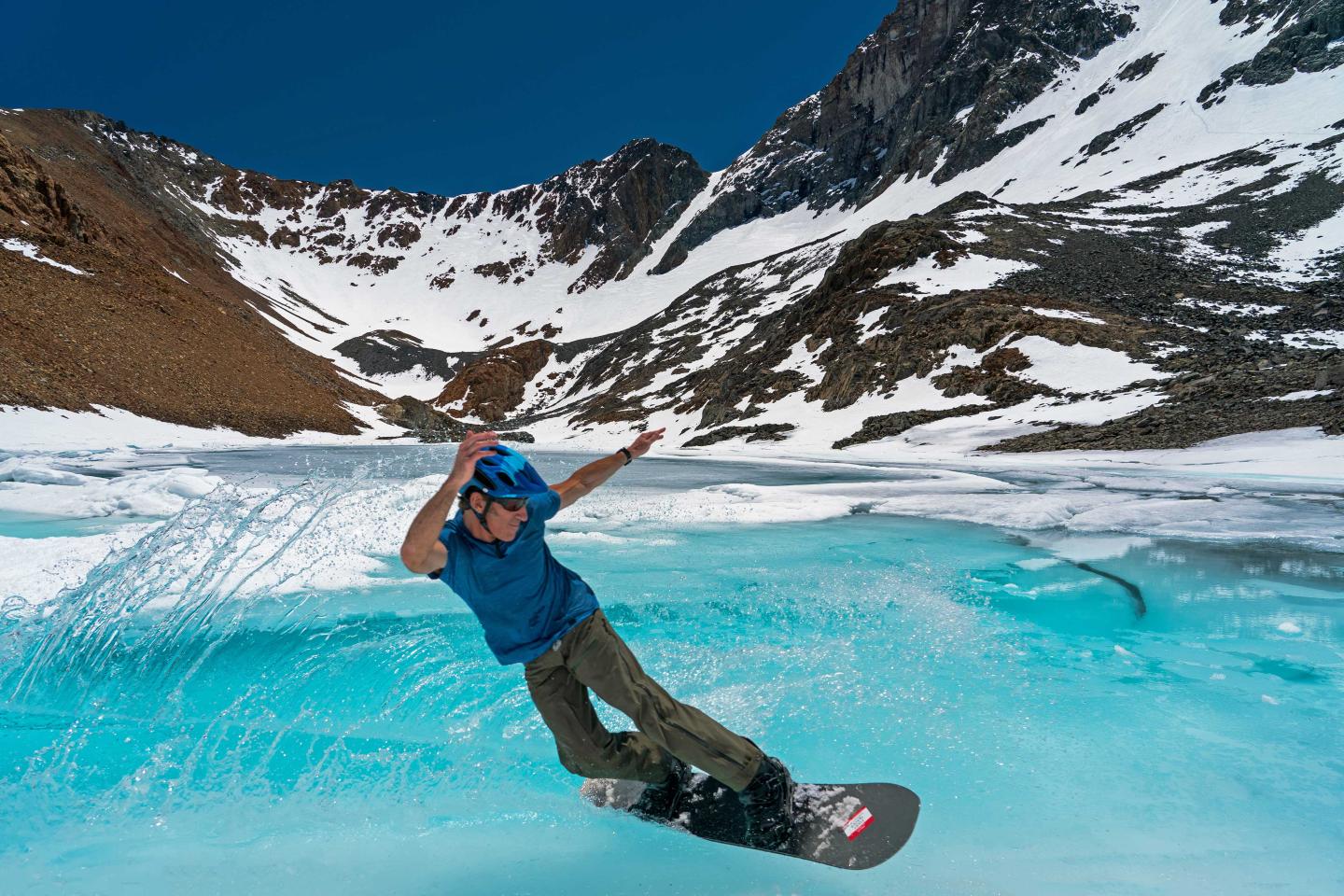 Snowboarder gliding over frozen turquoise lake amid snowy mountains.