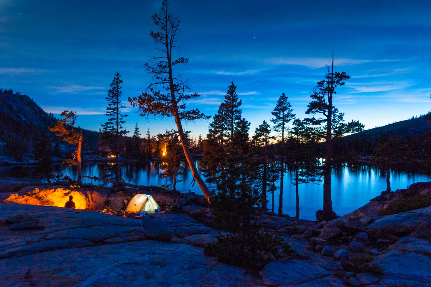 Camping by a lake at twilight, with tents lit and silhouetted trees.