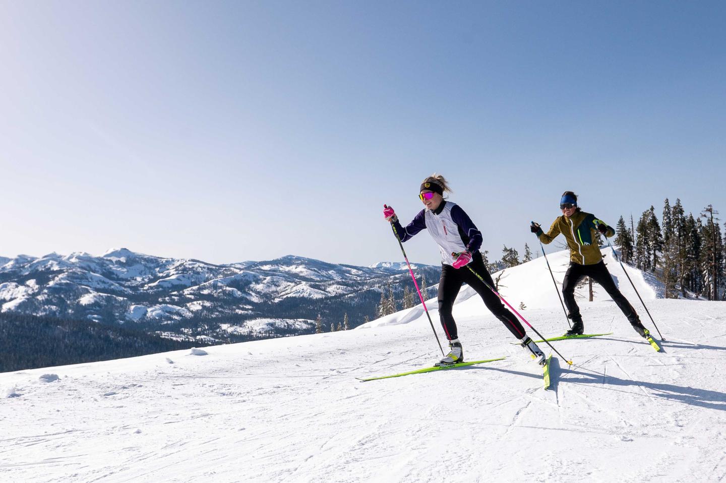 Two people cross-country skiing on a snowy mountain under a clear blue sky.