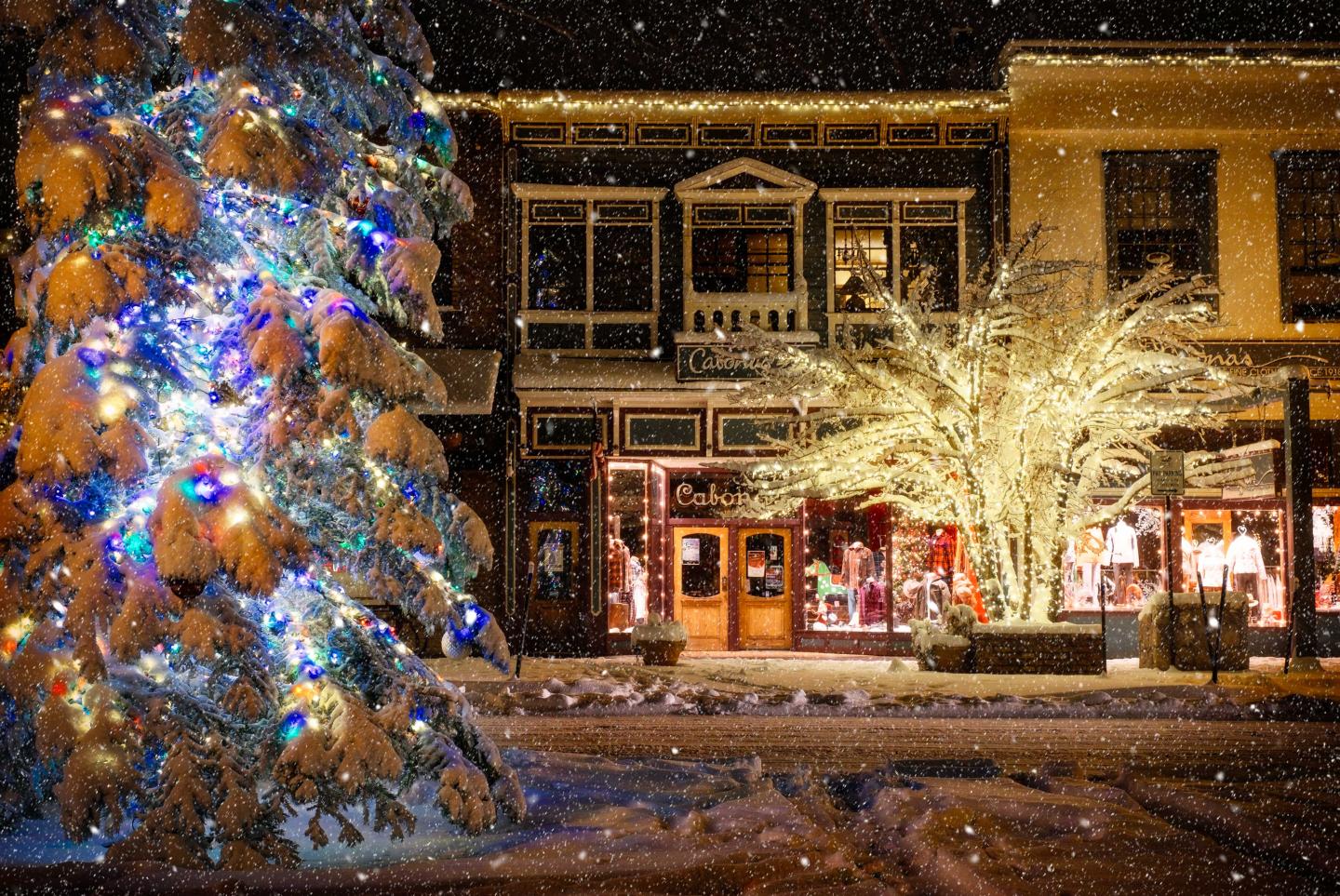 Snowy street with lit Christmas tree and festive storefronts at night.