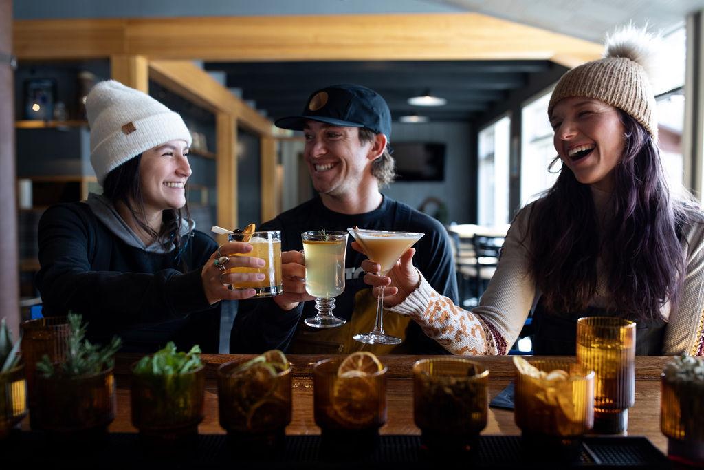 Three people in a cozy bar toasting with cocktails, smiling warmly.