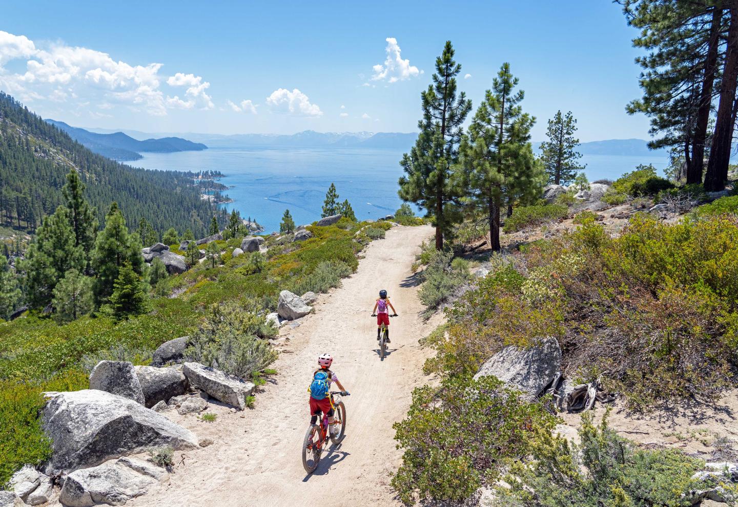Cyclists on a dirt path with lake and pine trees under a clear blue sky.
