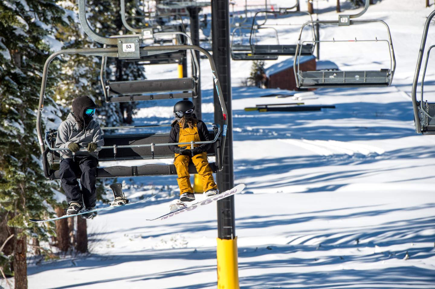 Two skiers on a snow-covered ski lift under a clear blue sky.