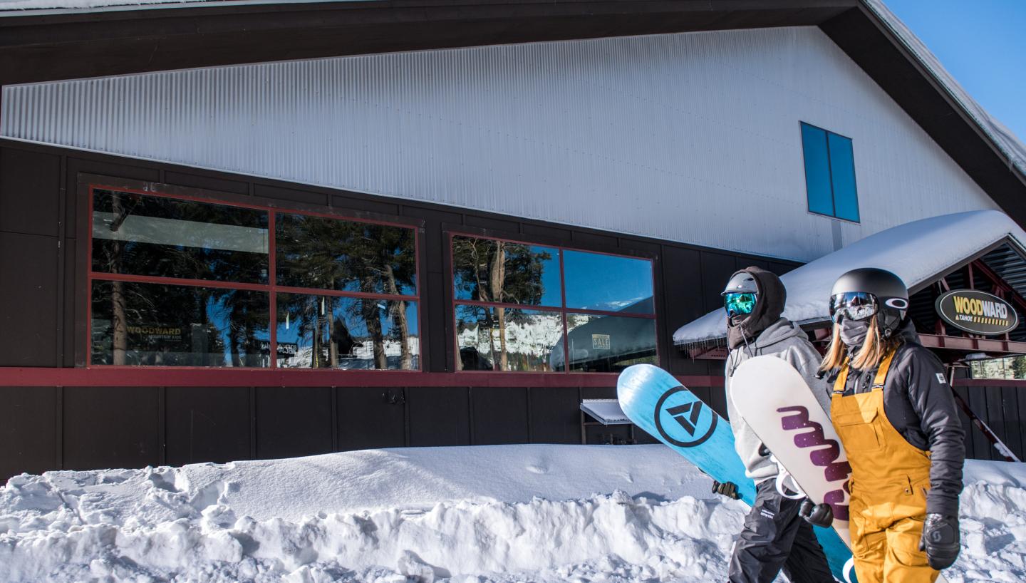 Snowboarders in gear walk past a snowy building under a clear sky.