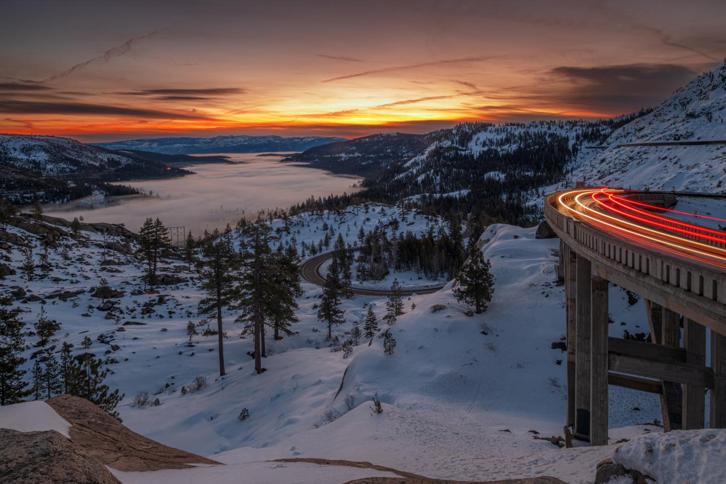 Snowy mountain road with light trails at sunset, overlooking a lake and distant hills.