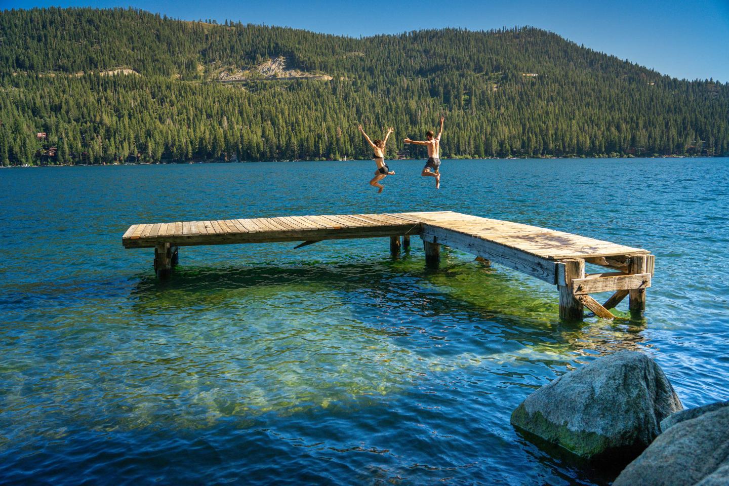 Two people jump off a wooden dock into a lake surrounded by trees and mountains.