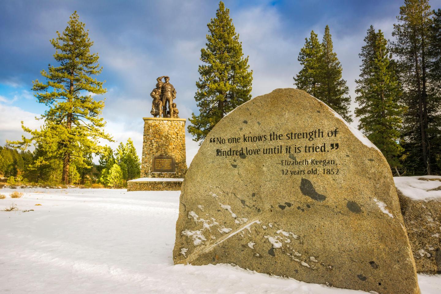 Donner Memorial Park / Stone monument and statue in a snowy landscape with pine trees under a blue sky.