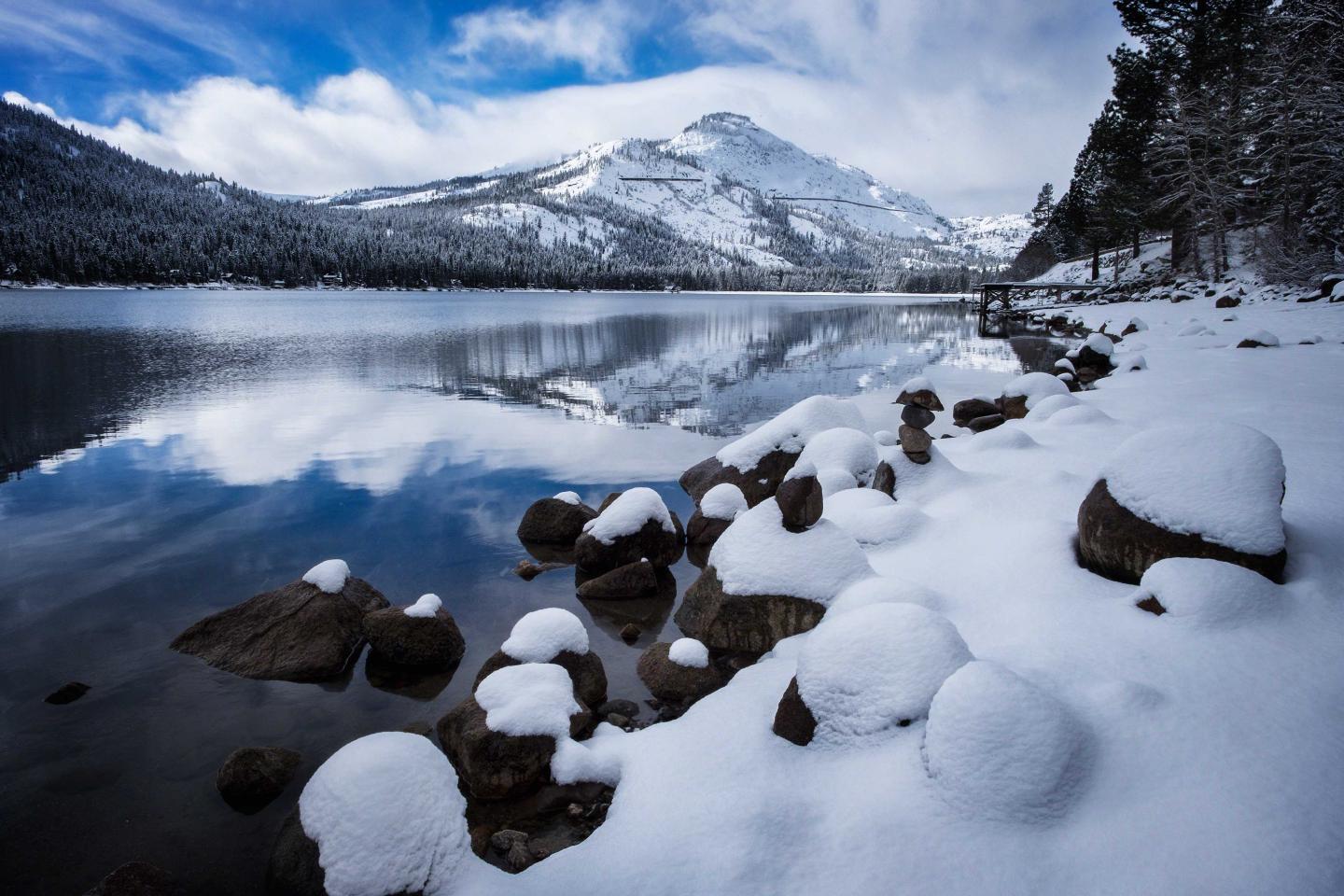 Snow-covered lakeside with distant mountains and blue sky reflections in water.