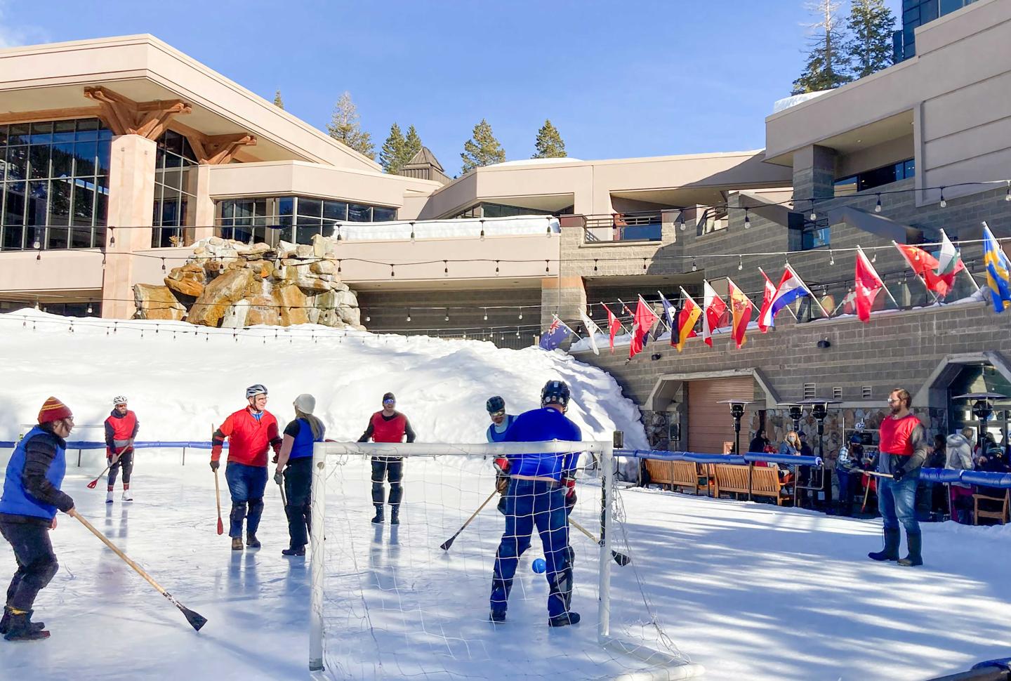 People playing ice hockey outdoors near buildings with flags and snowy surroundings.