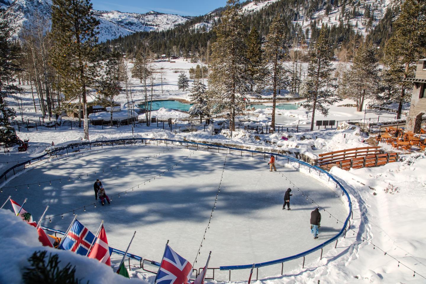 Snowy ice rink surrounded by flags, trees, and mountains in bright sunlight, Everline Resort, Olympic Valley, Palisades Tahoe
