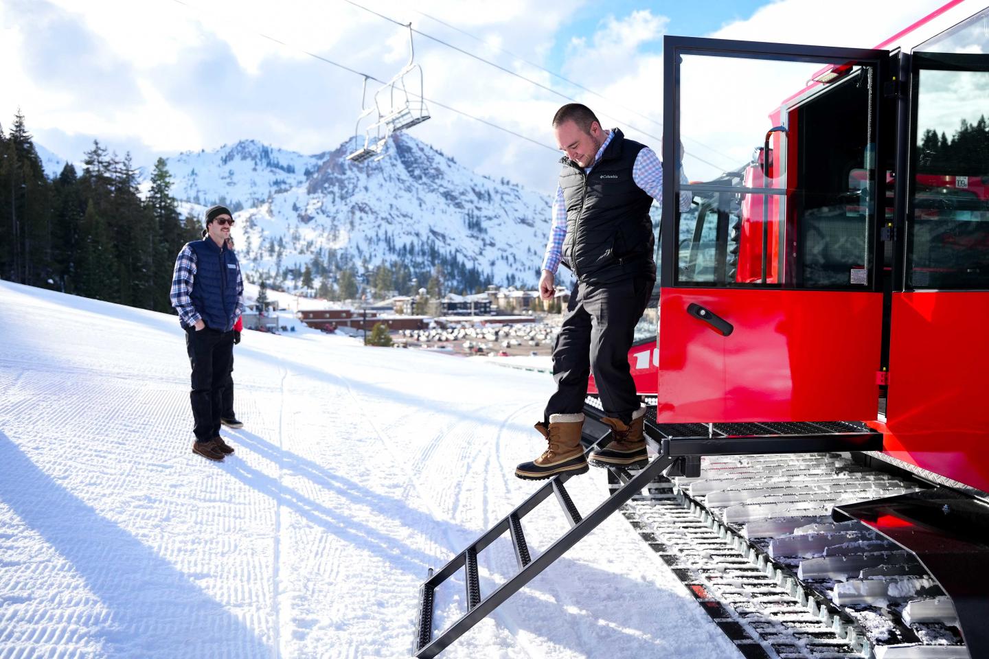 Man steps off red snowcat onto snowy mountain landscape; another man stands nearby.