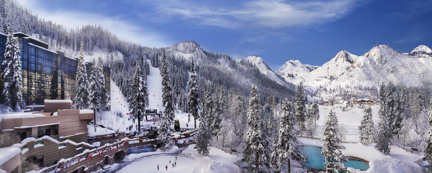 Snowy mountain resort with cabins, trees, a pool, and snow-covered hills under a blue sky.