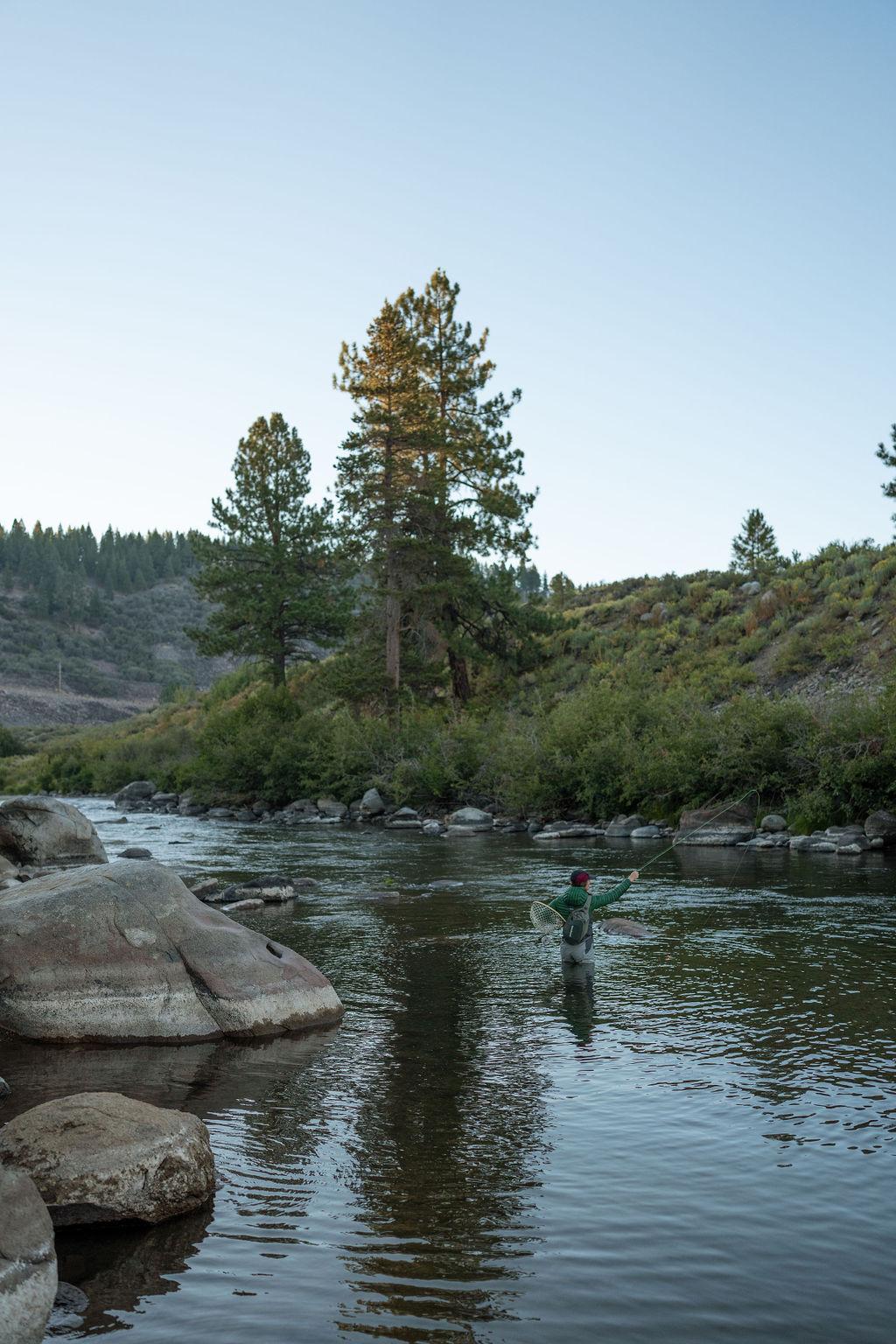 Person fly fishing in a calm river surrounded by trees and rocky banks.