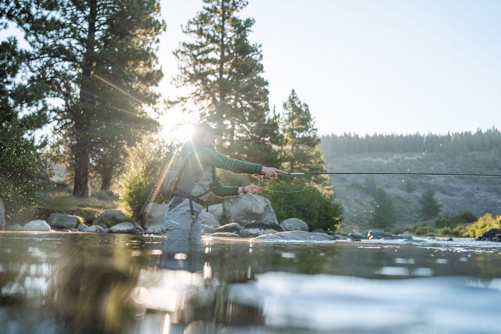 Person fly fishing in a river at sunrise, surrounded by trees.