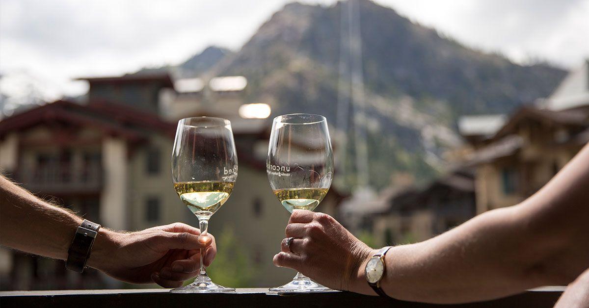 Two hands clink wine glasses on a balcony, with a mountain backdrop.
