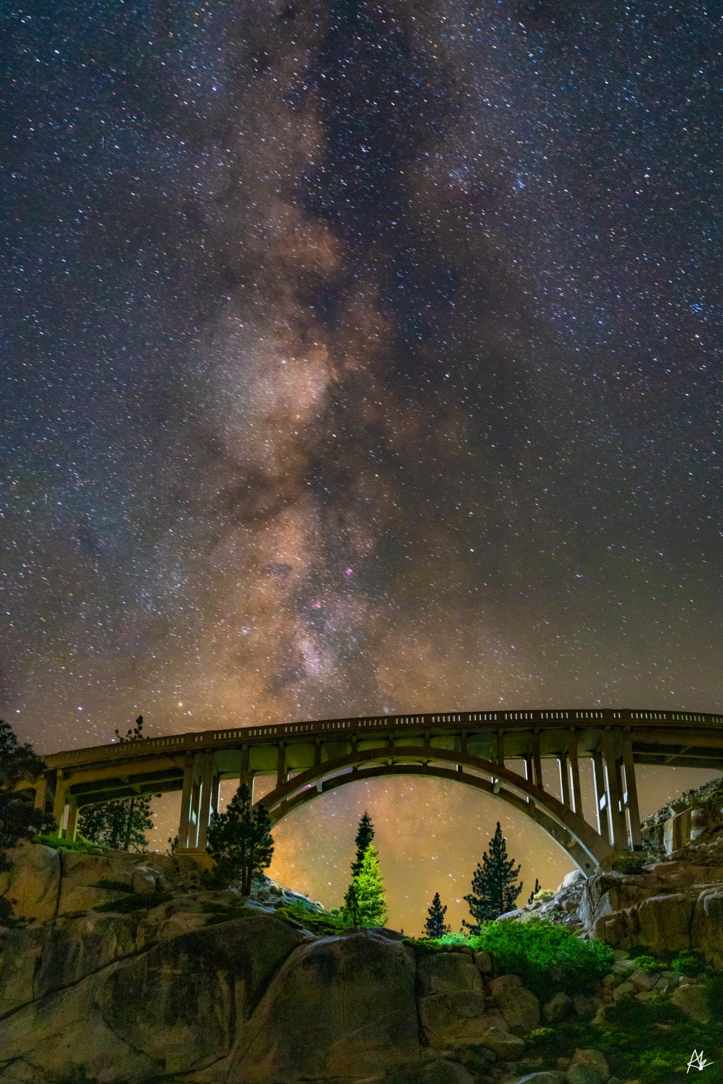 Starry night sky with Milky Way over a silhouetted bridge and trees.