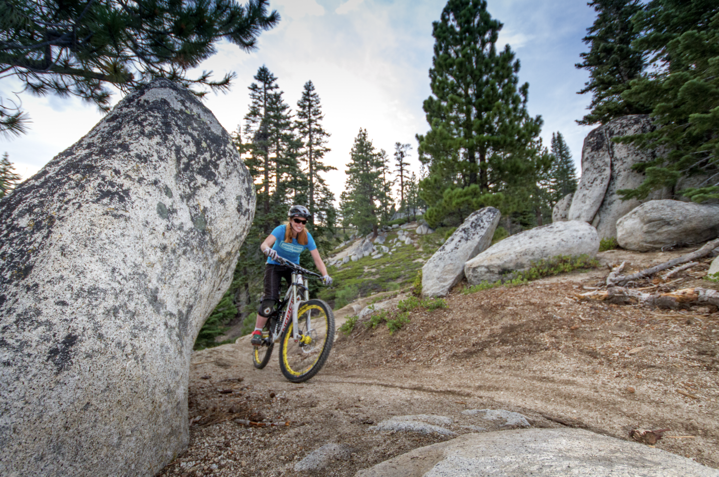 Cyclist rides through a forested rocky trail, surrounded by tall trees and large boulders.