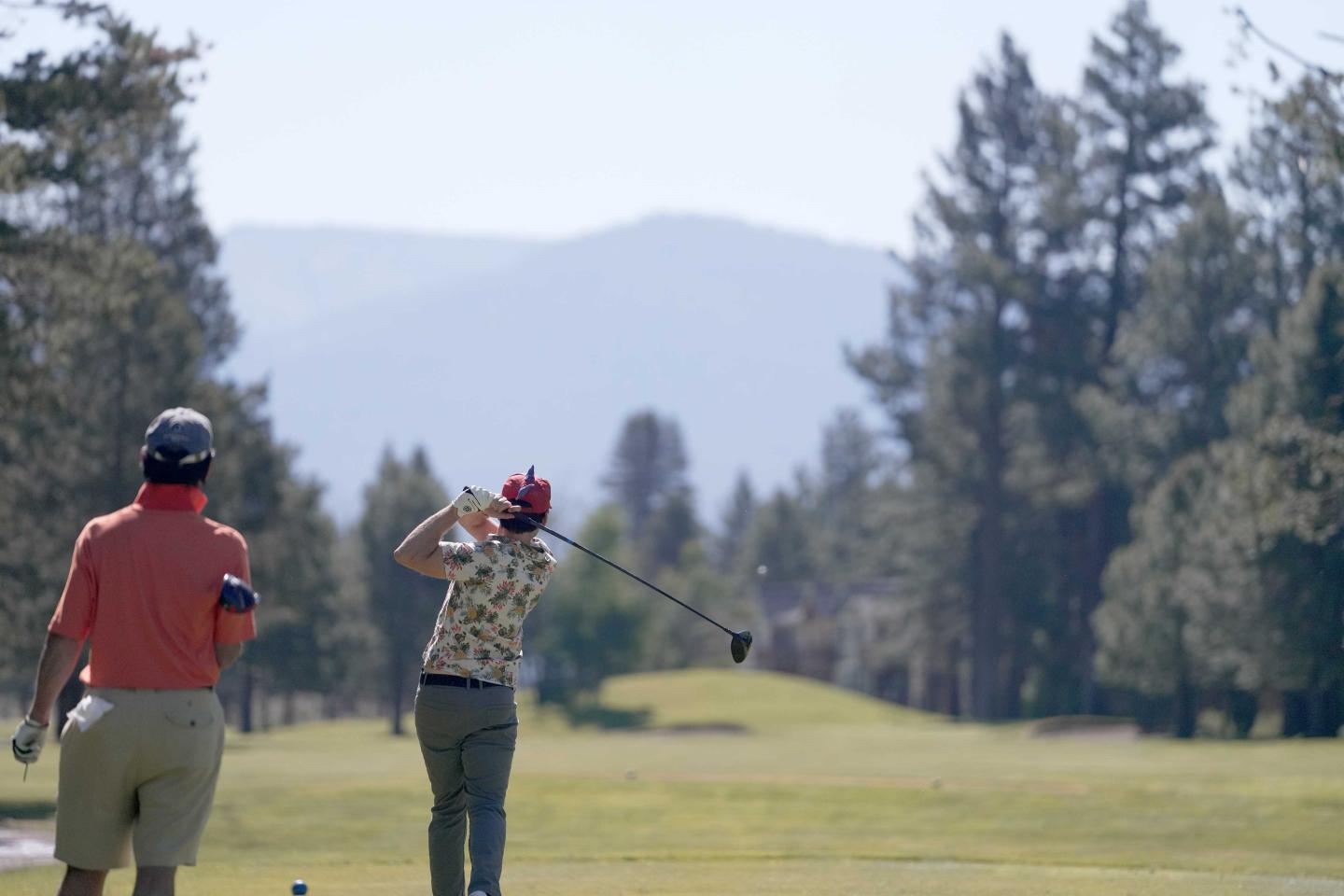Golfers on a course, one swinging, trees and mountains in the background.