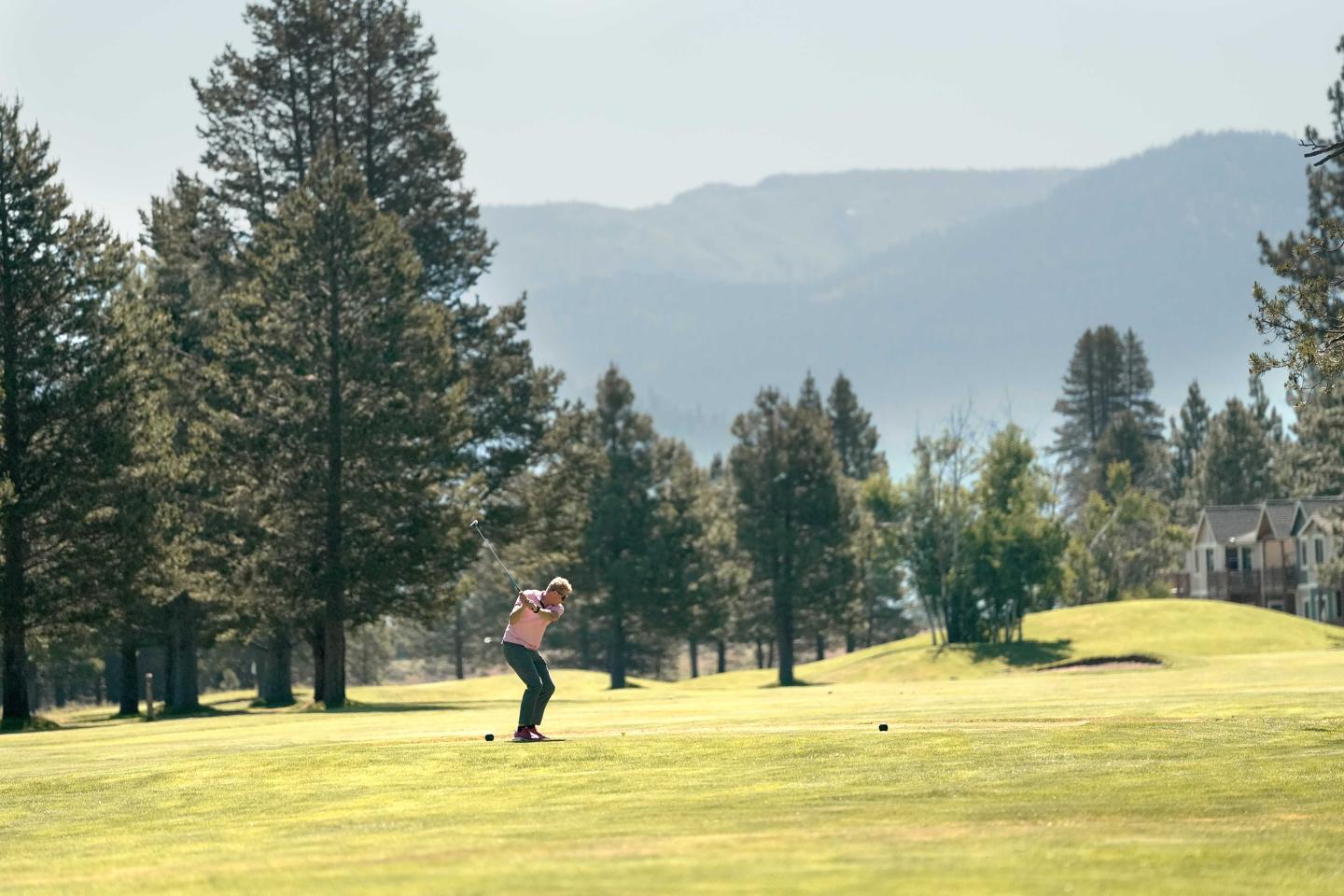 Golfer swinging on a green course with trees and mountains in the background.