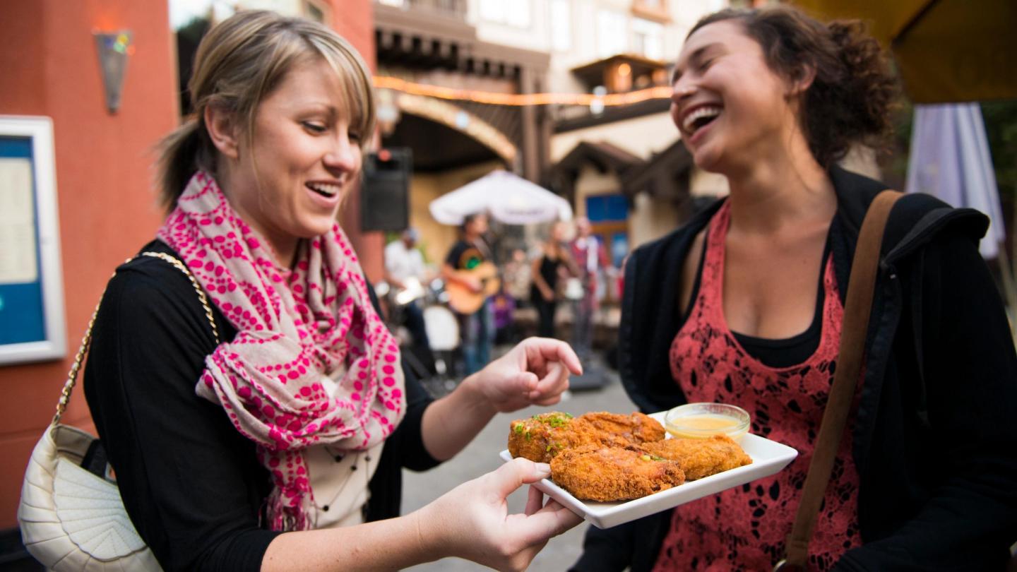 Guitar Strings vs. Chicken Wings / Two women laughing and sharing a tray of fried food outdoors.